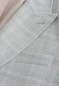 Revers de blazer à carreaux gris clair et poche poitrine sur fond de tissu beige, montrant les détails du motif tissé texturé.