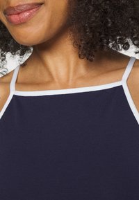 Marinefarbener Tanktop mit weißem Besatz, dünnen Trägern und geripptem Textil. Der Ausschnitt ist quadratisch und zeigt den Bereich des Schlüsselbeins.