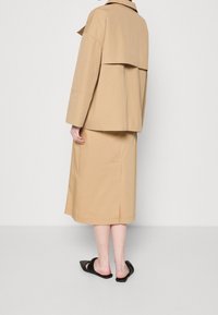 Beżowa, bawełniana kurtka oversize z opadającymi ramionami, warstwowym fasonem oraz dopasowaną spódnicą sięgającą kolan. Czarne, płaskie sandały na udach.