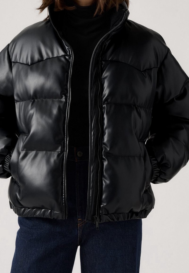 Personne portant une doudoune noire brillante par-dessus un col roulé noir et un jean bleu foncé, debout les mains sur les hanches.