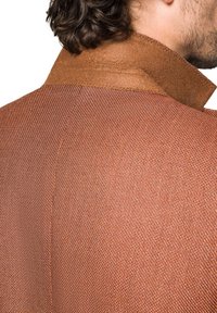 Brązowa kurtka o teksturowanej strukturze, z uniesionym kołnierzem. Tkanina ma pleciony wzór, uzupełniony gładkim kołnierzem w kontrastowym odcieniu brązu.