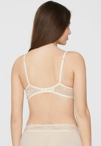 Sujetador beige con espalda de encaje, tirantes ajustables, borde festoneado y cierre de gancho y ojo. Textura de tela suave y patrón floral delicado.