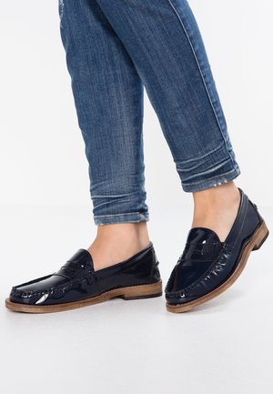 Mocassins en cuir verni bleu marine avec semelles en caoutchouc beige, dotés de coutures et d'un design à profil bas. Portés avec un jean bleu délavé.
