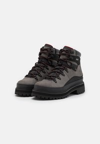 Harmaat vaelluskengät, joissa on korkea varsi, liian materiaali, mustat yksityiskohdat, tumman värinen kumipohja ja erottuvat oranssit nauhat; kestävä muotoilu ulkokäyttöön.