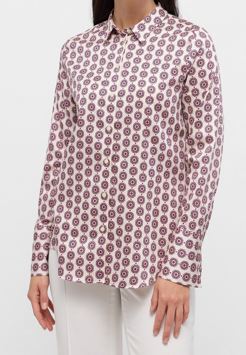 Femme portant une chemise à manches longues boutonnée avec un motif circulaire rose et noir, assortie à un pantalon blanc sur un fond uni.