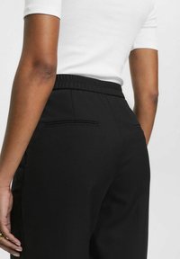 Pantalon noir avec une taille élastique, doté d'une texture lisse et d'un détail de poche arrière fine. Porté avec un haut blanc ajusté.
