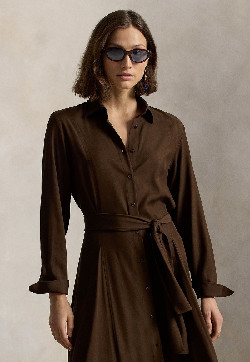 Γυναίκα που φορά ένα καφέ φόρεμα με μακριά μανίκια και κουμπιά, με ζώνη στη μέση δεμένη, σκούρα γυαλιά ηλίου και ένα κρεμαστό σκουλαρίκι, μπροστά σε ουδέτερο φόντο.