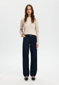 Maglione beige a maniche lunghe, abbinato a jeans in denim scuro a vita alta. I jeans hanno una vestibilità ampia e sono abbinati a una cintura nera.
