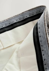 Graues und cremefarbenes Gewebe mit Fischgrätmuster, ausgestattet mit einer gewellten Kante, kontrastierendem schwarzen Besatz und glattem Innenfutter.
