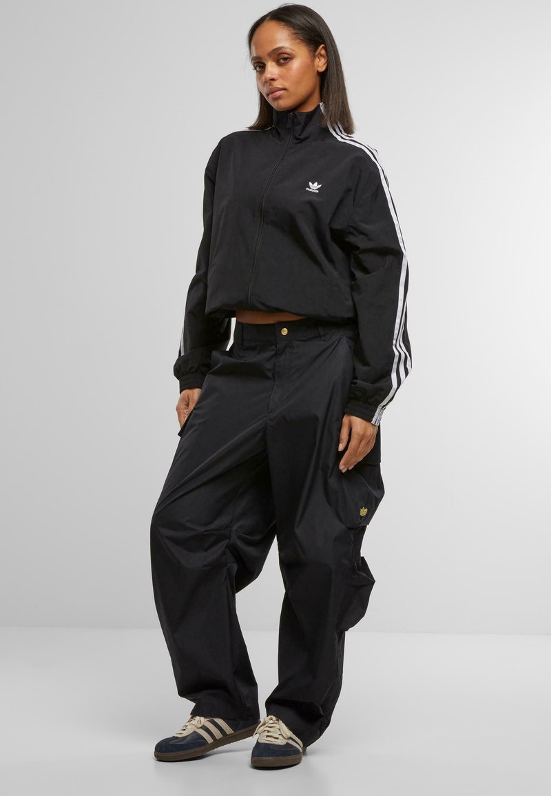 Schwarze Sportjacke mit weißen Streifen und Logo, kombiniert mit lockeren schwarzen Hosen mit Taschen und goldenen Akzenten. Blaue und cremefarbene Schuhe.