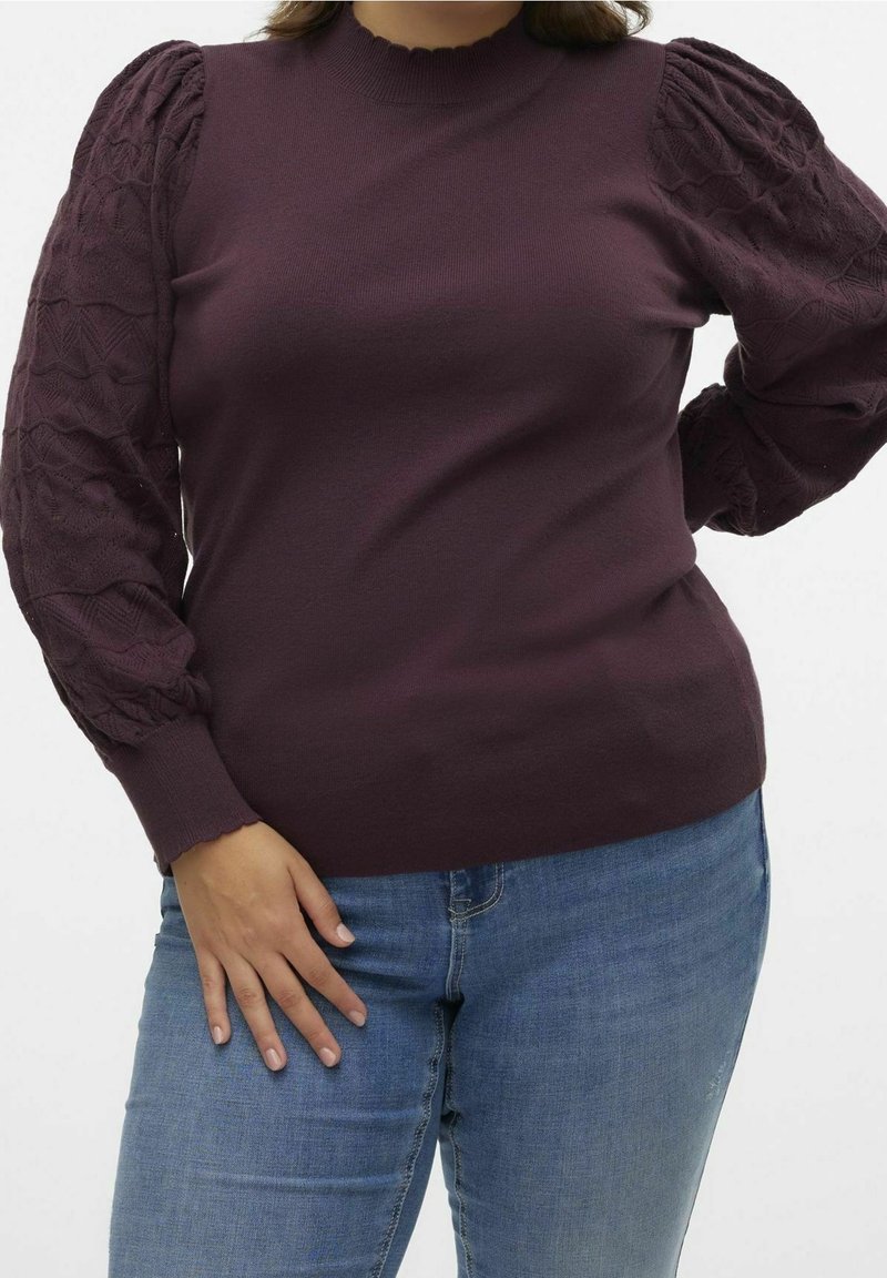 Donna che indossa un maglione di maglia testurizzata di un viola scuro con maniche a palloncino e jeans di denim blu, su uno sfondo neutro.