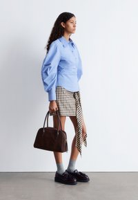 Light blue puff-sleeve shirt, checkered fabric skirt, brown suede handbag, gray socks, dark shoes, among a minimalist background.