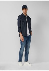 Granatowa koszula z długim rękawem z dwoma kieszeniami na piersi, biała koszulka pod spodem, niebieskie dżinsy i białe trampki. Uzupełniona pasującą granatową czapką.