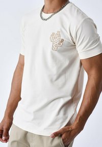 Crème t-shirt met korte mouwen met een textuur cactusborduring op de linkerborst en een ronde halslijn, gecombineerd met een zilveren ketting.