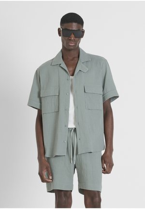 Camicia a maniche corte di un verde chiaro texture con due tasche sul petto, abbinata a pantaloni corti coordinati. Il modello indossa occhiali da sole neri di forma rettangolare.