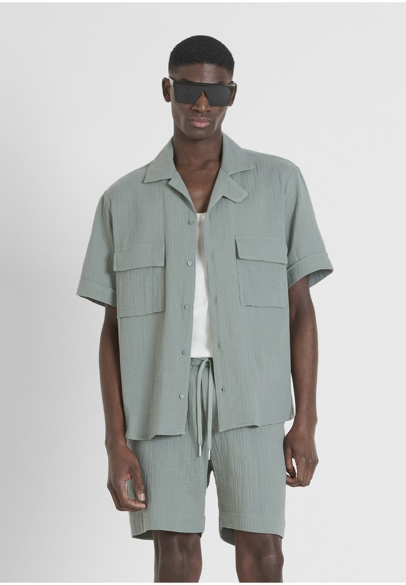 Lichtgroene gestructureerde shirt met korte mouwen en twee borstzakken, gecombineerd met bijpassende shorts. Het model draagt zwarte rechthoekige zonnebril.