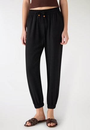 Donna che indossa pantaloni jogger neri con vita elastica, coulisse e polsini elastici, abbinati a sandali bassi marroni e top marrone corto.