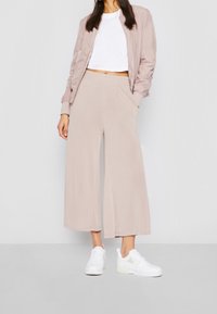 Veste bomber rose clair, t-shirt court blanc, pantalon large beige, et baskets blanches avec des accents transparents. Textures lisses, coupe décontractée.