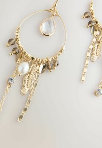 Gold hoop earrings featuring chains, leaf accents, a clear teardrop gemstone, and various brown and clear beads.