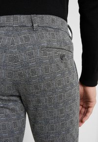 Person som bär grå rutiga byxor med en bakficka med knapp och en svart långärmad topp, hand vilande bredvid höften.