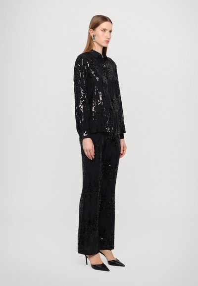 Musta paljettitakki ja housuasu, jossa on kauluspaita. Slim fit, sileä rakenne, korostettuna korkeakorkoisilla mustilla kengillä.