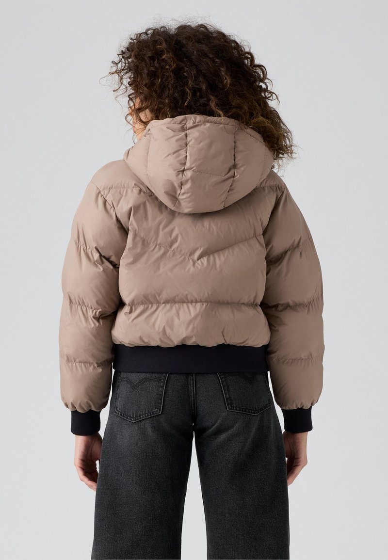 Giacca a vento beige, con cappuccio e polsini elasticizzati. Presenta un design trapuntato e una vestibilità corta. Indossata sopra jeans scuri.