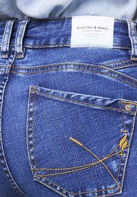 Blue denim jeans with a classic fit, featuring contrast yellow stitching and a unique embroidered design on the back pocket. Label visible.