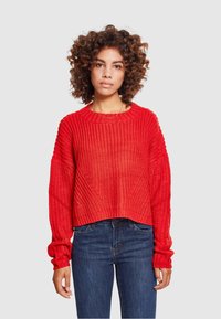 Kvinne med krøllete hår iført en lys rød strikkegenser og blå jeans som står foran en enkel lys grå bakgrunn.