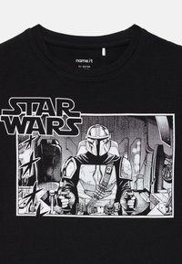 Svart T-skjorte med et hvitt grafikk av Star Wars' Mandalorian-karakter som pilot i et romskip, holder kontrollene inne i cockpiten.