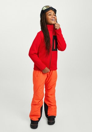 Giacca in pile rossa con una zip nera, pantaloni da neve arancioni e stivali neri. Modella che indossa occhiali da sci, sorridente con capelli intrecciati.