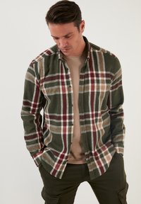 Camisa de franela en cuadros verdes y marrones, con botones, cuello y un corte relajado. Lleva una camiseta texturizada beige debajo.