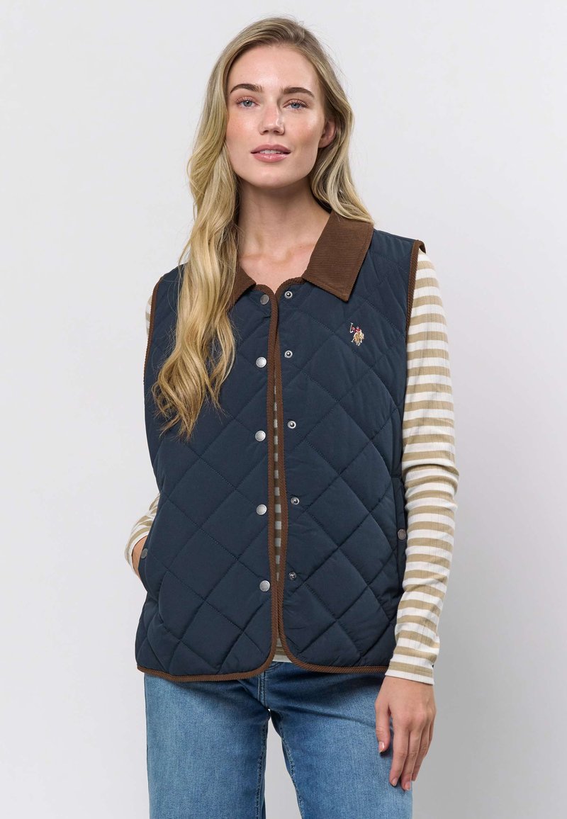 Quiltet marineblå vest med krage i brun kordfløyel, trykknappelukking og horisontal quiltesting. Har en brodert logo på brystet.