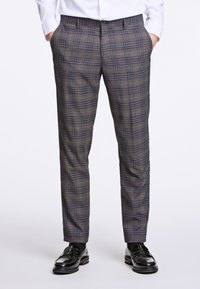 Pantaloni a quadri nei toni marrone e blu, realizzati in tessuto testurizzato, slim fit con tasche laterali e passanti per cintura. Abbinati a scarpe nere lucide.