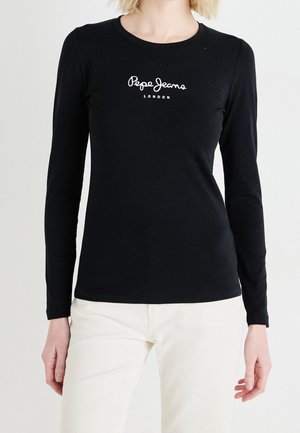 Kobieta ubrana w czarną koszulę z długim rękawem z logo "Pepe Jeans London" w połączeniu z białymi spodniami, stojąca na jednolitym tle.