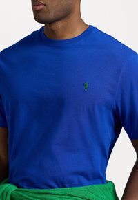 Královská modrá bavlněná tričko s kulatým výstřihem a krátkými rukávy. Na levé horní části hrudi je malá zelená vyšívaná logo.