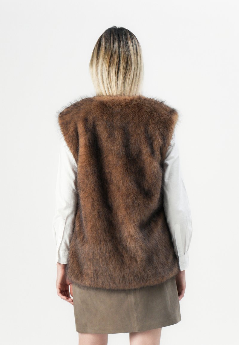 Persona con capelli biondi che indossa un gilet in pelliccia sintetica marrone su una camicia bianca a maniche lunghe e una gonna verde oliva, in piedi con le spalle rivolte verso l'osservatore su uno sfondo bianco.