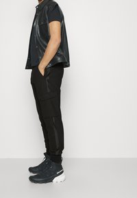 Veste sans manches en cuir noir sur un t-shirt noir, accompagnée d'un pantalon cargo noir avec des poches latérales et de chaussures de sport noires avec des accents blancs.