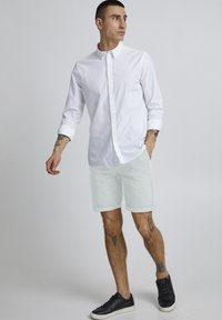 Camisa blanca de botones con mangas remangadas, pantalones cortos menta claros y zapatillas negras. Presenta tatuajes visibles en los brazos y las piernas.