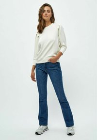 Sudadera blanca con mangas plisadas, combinada con jeans acampanados azules y zapatillas gris-blancas, sobre un fondo liso.