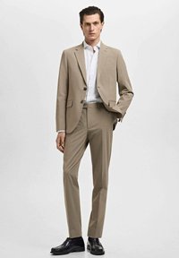 Traje beige a medida con una chaqueta ajustada, cierre de botón simple y pantalones a juego. Combinado con una camisa de vestir blanca y zapatos negros.