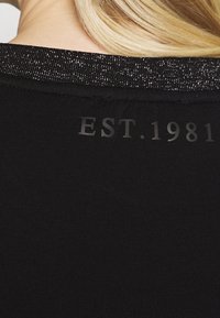 Czarna tkanina z teksturowanym, błyszczącym dekoltem. Na plecach znajduje się nadruk "EST. 1981" w ciemnoszarym kolorze. Prosty i nowoczesny design.