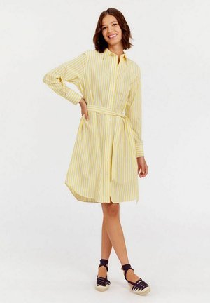 Robe chemise jaune à rayures verticales blanches, avec un boutonnage à l'avant, des manches longues et une ceinture assortie à la taille. Chaussures espadrilles.