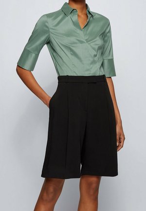 Kobieta ubrana w jasnozieloną koszulę na krótki rękaw, wpuszczoną w wysokie, czarne, do kolan, dopasowane szorty.