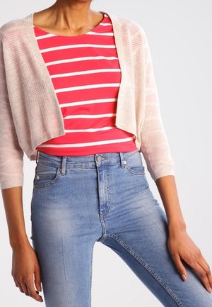 Persona con camiseta de rayas rojas y blancas, cárdigan corto rosa claro y vaqueros de tiro alto azul claro, de pie con las manos relajadas a los lados.