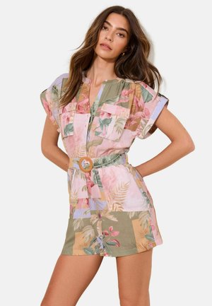 Žena, ktorá nosí krátky overal s pásikom a s pastelovým kvetinovým a listovým vzorom, stojí s rukami na bokoch na jednotnom pozadí.