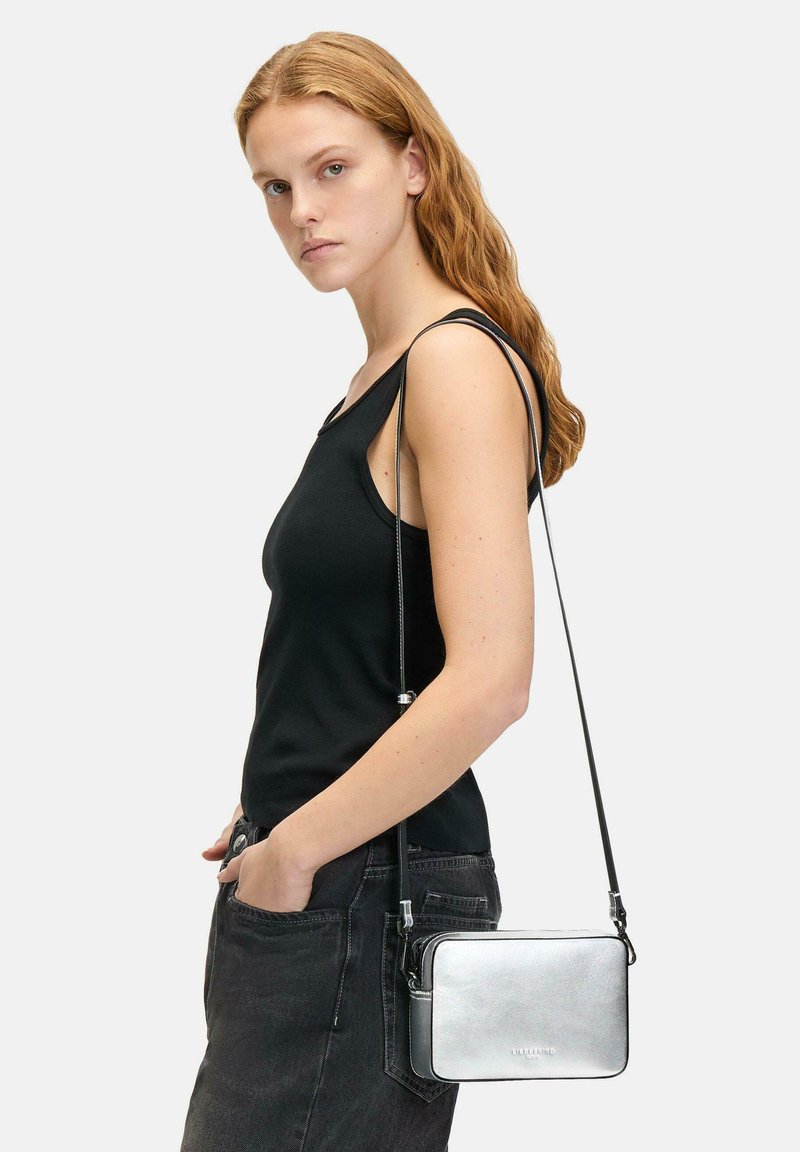 Silberne rechteckige Umhängetasche mit strukturiertem Finish, schwarzem Schultergurt und Reißverschluss, getragen zu einem schwarzen Tanktop und dunklen Jeans.