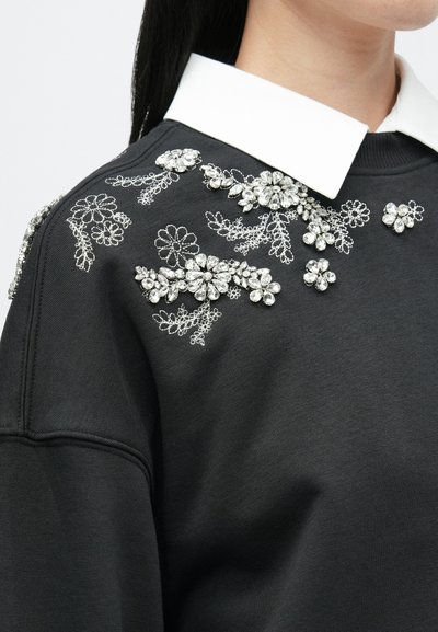 Μαύρη μπλούζα με λευκό γιακά, διακοσμημένη με περίπλοκα ασημένια λουλούδια κεντήματος και κρυστάλλινες λεπτομέρειες στον αριστερό ώμο.