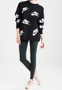 Felpa nera con loghi bianchi Nike, abbinata a leggings verde scuro e scarpe da ginnastica rosa chiaro. Texture lisce, vestibilità comoda.