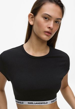 Fiatal nő egyenes barna hajjal, fekete crop topot visel, melyen a deréknál fehér, Karl Lagerfeld márkajelzésű rugalmas szalag található.