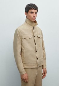 Beige Jacke mit hohem Kragen, zwei Brusttaschen, Knopfverschluss vorne und strukturiertem Stoff. Das Modell trägt beigen Hosen im minimalistischen Design.